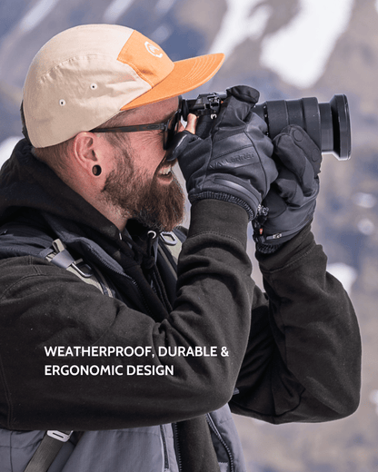 A man wearing a beige and orange baseball cap, black Vallerret Markhof Prov4 gloves, and a dark jacket looks through a camera lens outdoors with snowy mountains in the background. White text at the bottom reads: &